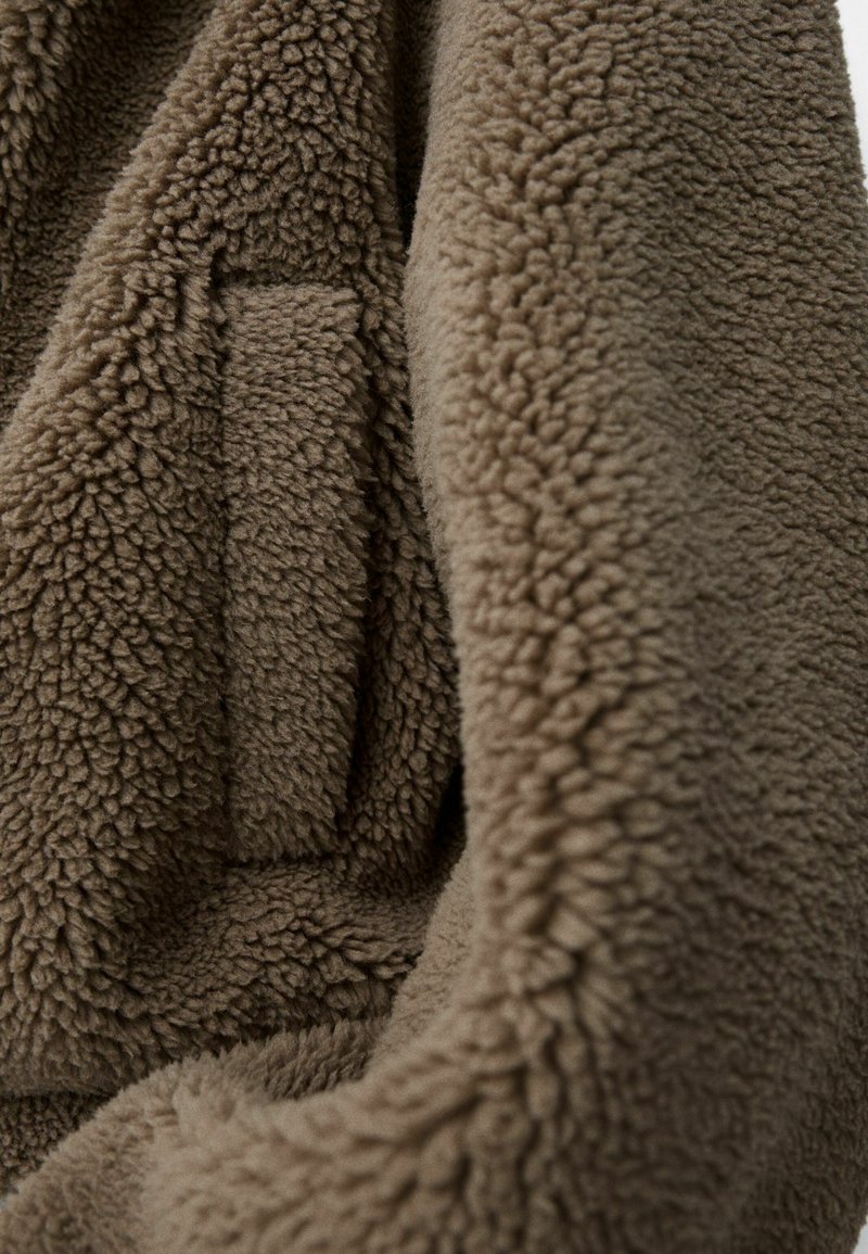 Tissu en polaire marron avec une surface texturée, présentant un détail de poche et une apparence douce et duveteuse, tombant en plis.