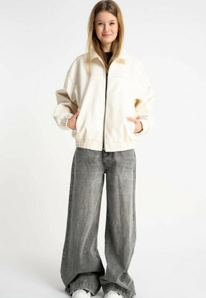 Młoda kobieta o długich włosach, ubrana w kremową kurtkę zapinaną na zamek, szerokie szare jeansy i casualowe buty, stojąca z rękami w kieszeniach.