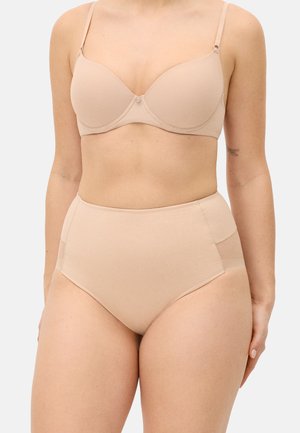 Mujer con un sujetador sin costuras en tono nude, con aros y relleno, y ropa interior de talle alto sin costuras contra un fondo claro y liso.