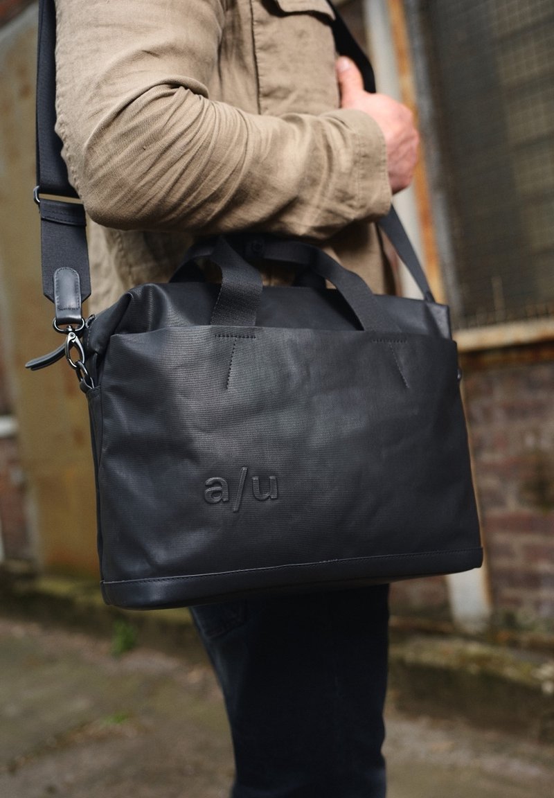 Homme portant une veste beige porte un sac à bandoulière noir avec "a/u" embossé sur le devant, debout près d'un mur en briques et d'une fenêtre.