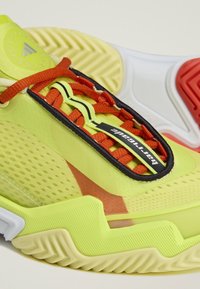 Close-up van een geelgroene sportschoen met oranje veters en een dikke zool, met een "Barricade" label op de tong en het Adidas-logo.