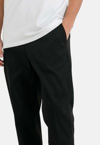 Zwarte broek met een relaxte pasvorm, voorzien van zijzakken, een gladde textuur en een licht kreukelige uitstraling. Gecombineerd met een witte t-shirt.