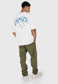 Hvid bomulds t-shirt med blå grafisk tekst på ryggen. Et par olivengrønne cargo-bukser med sidelommer, båret med lyse sneakers.