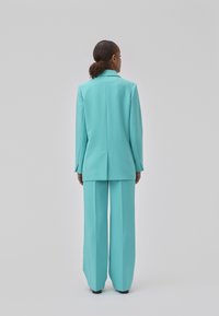 Een turquoise pak met een getailleerd colbert met insnoerde revers en brede broekspijpen, gemaakt van een soepele stof, gezien vanaf de achterkant.