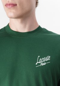 T-shirt en coton vert avec col rond et logo brodé "Lacoste Paris" blanc sur la poitrine gauche. Texture lisse, coupe décontractée.