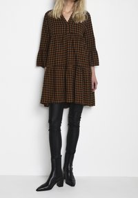Braun- und schwarz kariertes Kleid mit einem gestuften Design und drei Viertel Ärmeln, kombiniert mit eng anliegenden schwarzen Lederhosen und spitzen Blockabsatzstiefeln.