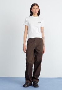 Camiseta branca de mangas curtas com a marca "Dickies", combinada com calças largas castanhas e ténis pretos sobre um chão de cor clara.