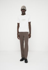 Balti marškinėliai su juodu logotipu, rudi siūti kelnės ir smėlio spalvos kepurė. Juodi batai užbaigia aprangą. Minimalistinis dizainas ir neutralios spalvos.