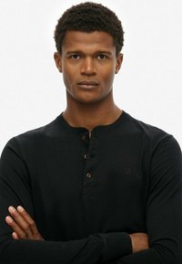 Zwarte langemouwen henley-shirt gemaakt van zachte stof, met een ronde halslijn, drie bruine knopen en een klein logo op de borst.