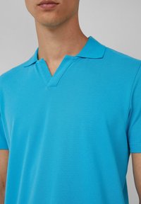 Koszulka polo z krótkim rękawem w jaskrawym turkusie, z dekoltem w serek i fakturowanym materiałem. Zbliżenie na kołnierz i górną część klatki piersiowej.