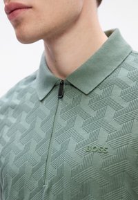 Gros plan sur une personne portant un polo vert à motifs avec une fermeture éclair et le logo "BOSS" sur la poitrine.
