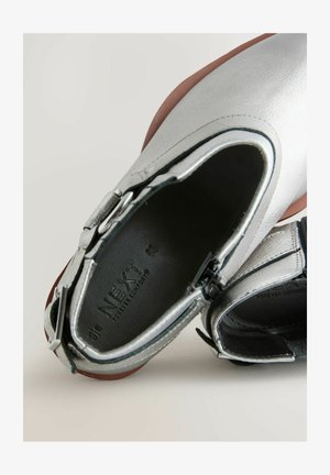 Srebrni usnjeni gležnjarji z črnimi detajli, z gladko teksturo, zaobljenim nosom in zadrgo. Notranjost prikazuje blagovno znamko in rjavo podplat.