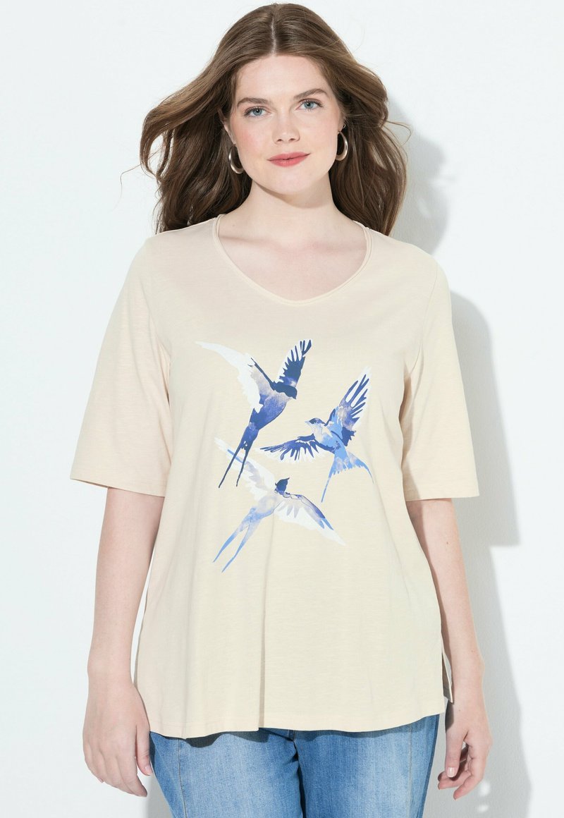 Mujer con cabello largo castaño, viste una camiseta beige con estampado de pájaro azul y jeans azules, de pie frente a un fondo blanco.