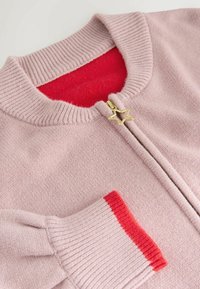 Světle růžový zip-up kardigan s červenou podšívkou. Vybavený ribbed límcem a manžetami, zlatým hvězdicovým táhlem zipu a texturovanou látkou.