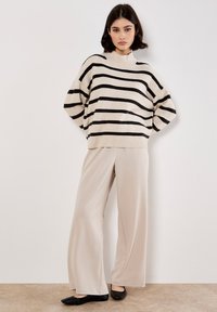 Maglione lavorato a maglia color crema con strisce nere, maniche larghe e colletto alto, abbinato a pantaloni beige fluidi e scarpe nere slip-on.