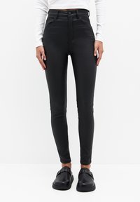 Jean skinny noir taille haute en matière lisse et extensible. Comprend des poches avant et des détails de coutures discrètes. Porté avec des chaussures slip-on noires.