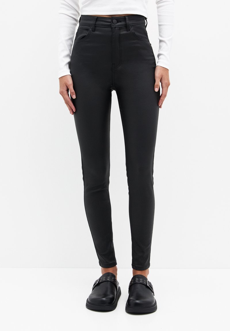 Jean skinny noir taille haute en matière lisse et extensible. Comprend des poches avant et des détails de coutures discrètes. Porté avec des chaussures slip-on noires.