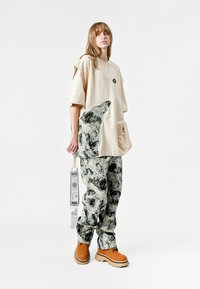 Persona joven vistiendo una camiseta beige de gran tamaño con dobladillo irregular y pantalones a juego con un patrón abstracto, sosteniendo una etiqueta, de pie contra un fondo blanco.