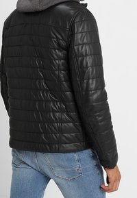 Man som bär en svart kviltad läderjacka över en grå hoodie och ljusblå jeans, visad bakifrån mot en enfärgad bakgrund.