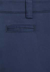 Granatowy materiał spodni z płaską tylnią kieszenią, subtelnymi detalami przeszycia i gładką teksturą, podkreślający dopasowany fason.