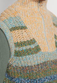 Zbliżenie na wielokolorową dzianinową kamizelkę na zamek noszoną na oliwkowo-zielonej, prążkowanej, długorękawowej koszuli.