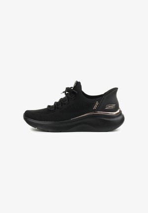 Scarpa sportiva nera con parte superiore in rete, punta arrotondata, suola testurizzata e un accento dorato lucido sul tallone. Presenta lacci standard.