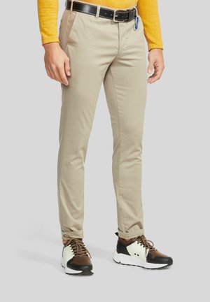 Man draagt beige chino's, zwarte riem, bruine en witte sneakers en een gele longsleeve, staand op een lichtgrijze achtergrond.
