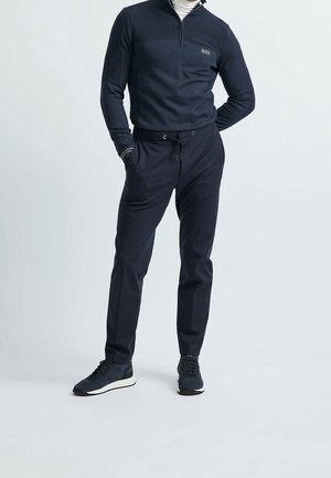 Man wearing navy blue zip-up sweater, dark drawstring pants, and navy sneakers, standing with hands in pockets against plain background.