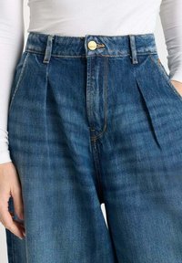 Pantalones de mezclilla azul con una cintura alta, que presentan pliegues y un cierre con botón dorado. Textura suave con desvanecimientos sutiles y costuras tradicionales.