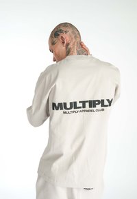 Světle béžová košile s dlouhým rukávem, na zádech a rukávech s černým textem "MULTIPLY". Měkká textura, volný střih, kulatý výstřih a spadlé ramena.