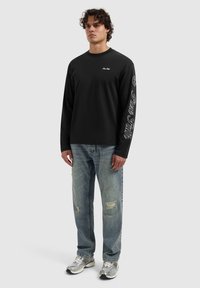 Zwarte lange mouwen shirt met wit logo; lichtblauwe versleten jeans; zilveren sneakers met gaasaccenten.