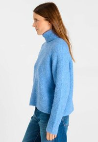 Vrouw met lang bruin haar, gekleed in een fluffy lichtblauwe turtlenecktrui en blauwe spijkerbroek, die naar links kijkt tegen een effen witte achtergrond.