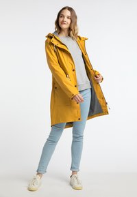 Gul vattentät jacka med huva, framdragkedja och två sidofickor. Bärs över en grå skjorta, i kombination med ljusblå jeans och vita sneakers.