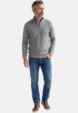 Man with gray hair and beard wearing a gray quarter-zip sweater, blue jeans, and brown leather sneakers standing with one hand in pocket.