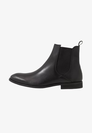 Botte Chelsea en cuir noir avec panneaux élastiques latéraux, finition lisse, bout rond et petit talon bloc. Comprend une languette à l'arrière.