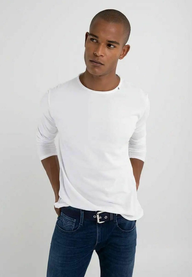 Camicia bianca a maniche lunghe con scollatura circolare, design aderente e dettaglio di branding discreto sul petto, abbinata a jeans blu scuro.