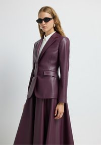 Blazer en cuir bordeaux avec coupe ajustée, revers et poches avant. Associé à une jupe assortie, avec une texture lisse.