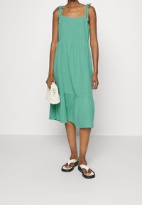 En knälång klänning i ljus teal med tank-style, med justerbara band och en flikig nederdel. Matchas med svartvita flip-flops.