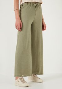 Lichte olijfgroene wijde broek van een textuurstof, met een verstelbare taille en een ontspannen pasvorm, gecombineerd met beige sneakers.