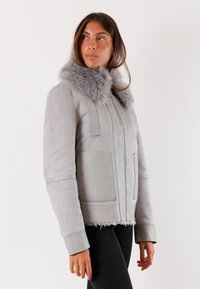 Giacca in suede grigia con colletto peloso, chiusura con zip, due tasche frontali e maniche larghe. Tessuto morbido con dettagli lisci e soffici.