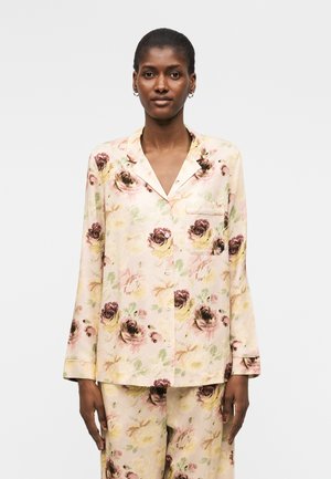 Moteris dėvi kreminės spalvos pižamos komplektą su rožiniu, geltonu ir rudu gėlių raštu, stovinti šviesiai pilkoje fono.