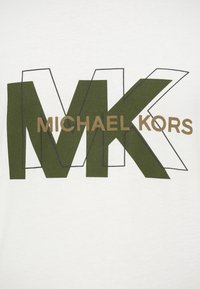 Michael Kors logo ar lieliem olīvzaļiem "MK" burtiem un mazākiem brūniem "MICHAEL KORS" teksta burtiem, kas pārklājas uz baltas auduma fona.