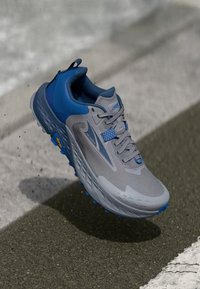 Grijze sportschoen met blauwe accenten, bovenwerk van mesh, gestructureerde zool en reflecterende details, ontworpen voor grip en stabiliteit.
