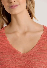 Eine Frau mit langen blonden Haaren trägt ein korallenrotes, gestricktes V-Ausschnitt-Oberteil, das ihren Hals und den unteren Teil ihres Gesichts zeigt, mit einem sanften Lächeln.