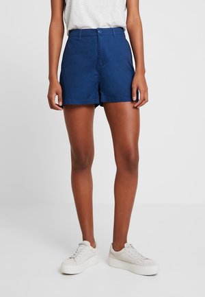 Femme portant un short taille haute bleu marine, un t-shirt blanc rentré, et des baskets blanches à plateforme, debout devant un fond clair uni.