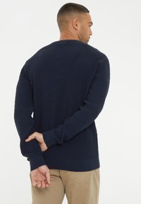 Marineblauer Strickpullover mit strukturiertem Rautenmuster, gerippten Bündchen und Saum. Rückwärts getragen, wodurch die Designmerkmale auf der Rückseite sichtbar sind.