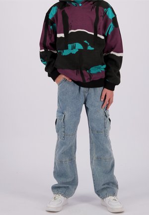 Sweat à capuche graphique violet vif et bleu sarcelle avec des accents noirs, associé à un jean cargo ample bleu clair et des baskets blanches. Capuche ajustable.