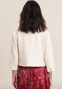 Afgekorte crèmekleurige blazer met een gladde textuur en driekwartmouwen. Heeft een eenvoudig ontwerp en een subtiel knopdetail bij de manchetten.