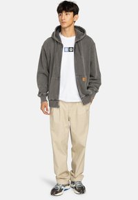 Sudadera gris con cremallera y gran bolsillo frontal, combinada con una camiseta blanca, pantalones beige y zapatillas plateadas. La sudadera cuenta con cordones ajustables.