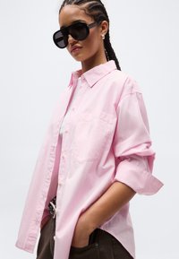 Γυναίκα με πλεξούδες, φοράει μεγάλα μαύρα γυαλιά ηλίου, ανοιχτό ροζ πουκάμισο με κουμπιά και μανίκια γυρισμένα και σκούρο παντελόνι με τα χέρια στις τσέπες.